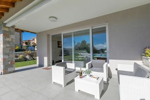 Eine überdachte Terrasse mit weißen Korbmöbeln, einer Glastür, die zu einem Pool führt, und einer Steinpfeiler.