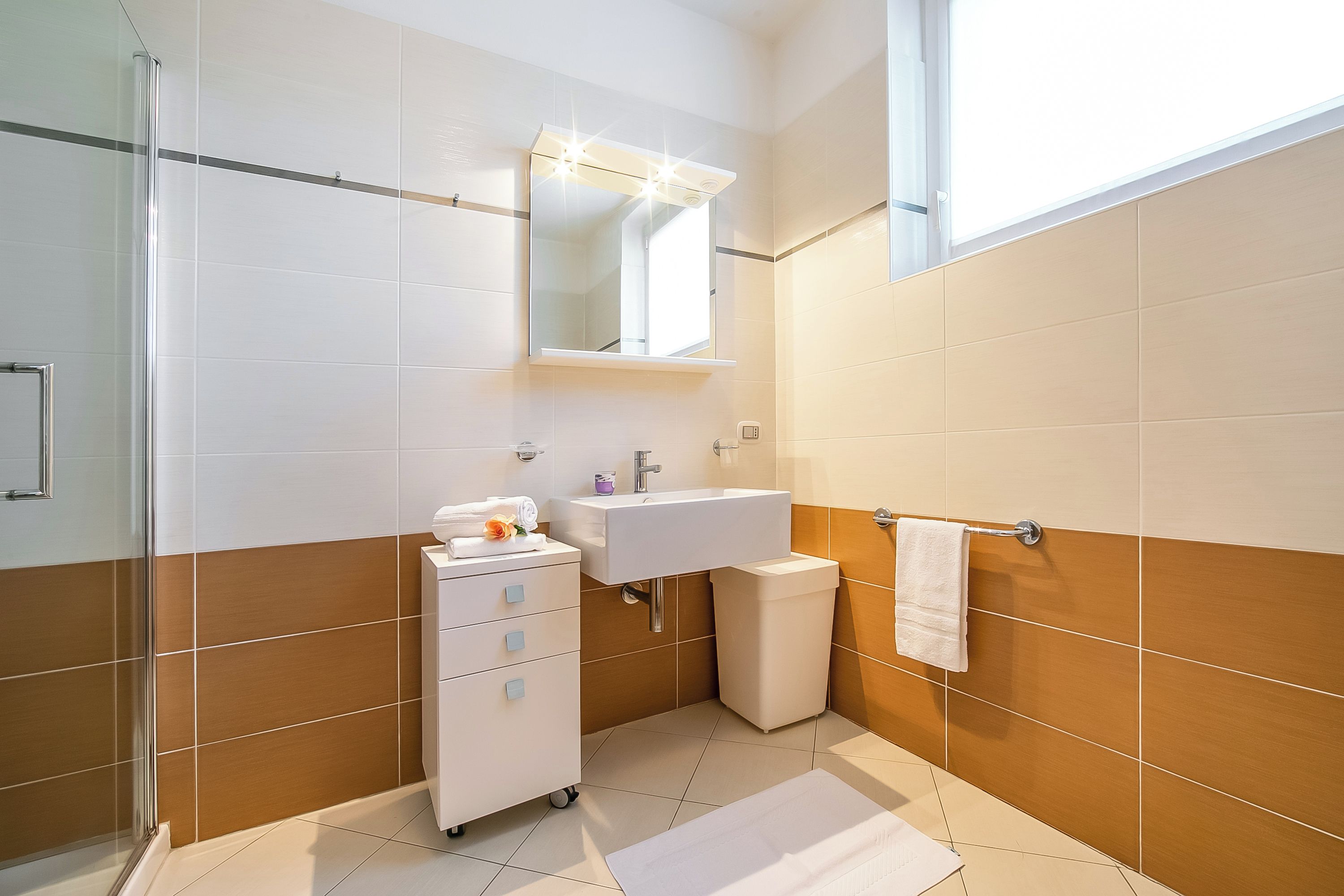 A bathroom with a glass shower enclosure, a white sink, a mirror with lights, and a towel rack.