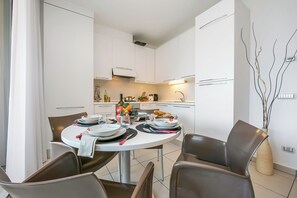 A modern kitchen with white cabinets, a dining table set for two, and a vase with branches on the wall. - Vista di Maccagno Fantastico Pool (Maccagno con Pino e Veddasca)