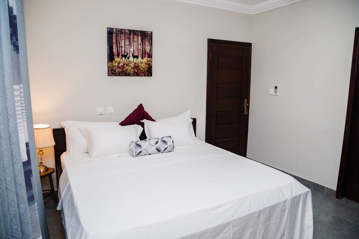 A bedroom with a bed, a nightstand, a lamp, a door, and a painting on the wall.