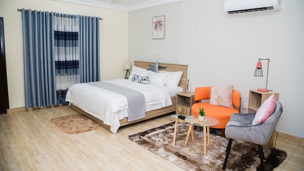 A modern hotel room with a bed, a small table with a cactus, a chair, a lamp, and an air conditioning unit. - Kasa Nautica Homes & Comfort (Accra)