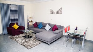A living room with a grey sectional sofa, a red armchair, a glass coffee table, a patterned rug, and a dining area with a glass table and metal chairs. - Kasa Nautica Homes & Comfort (Accra)