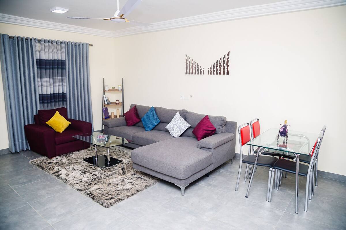 A living room with a grey sectional sofa, a red armchair, a glass coffee table, a patterned rug, and a dining area with a glass table and metal chairs.