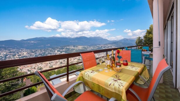 Uma varanda com mesa posta para refeições, com vista para a paisagem urbana e montanhas.