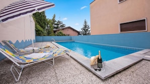Piscina com espreguiçadeiras e um guarda-sol, cercada por um edifício com fachada azul e bege.