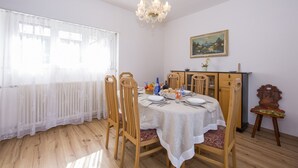 Uma área de refeições com mesa posta para quatro, cadeiras de madeira, um candelabro, um quadro na parede e um radiador sob uma cortina transparente.
