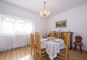A dining area with a table set for four, wooden chairs, a chandelier, a painting on the wall, and a radiator under a sheer curtain. - Aldesago Panoramica (Lugano)