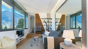 Un salon moderne avec un canapé, une table basse et une lampe. Une grande fenêtre offre une vue sur les montagnes et la ville.