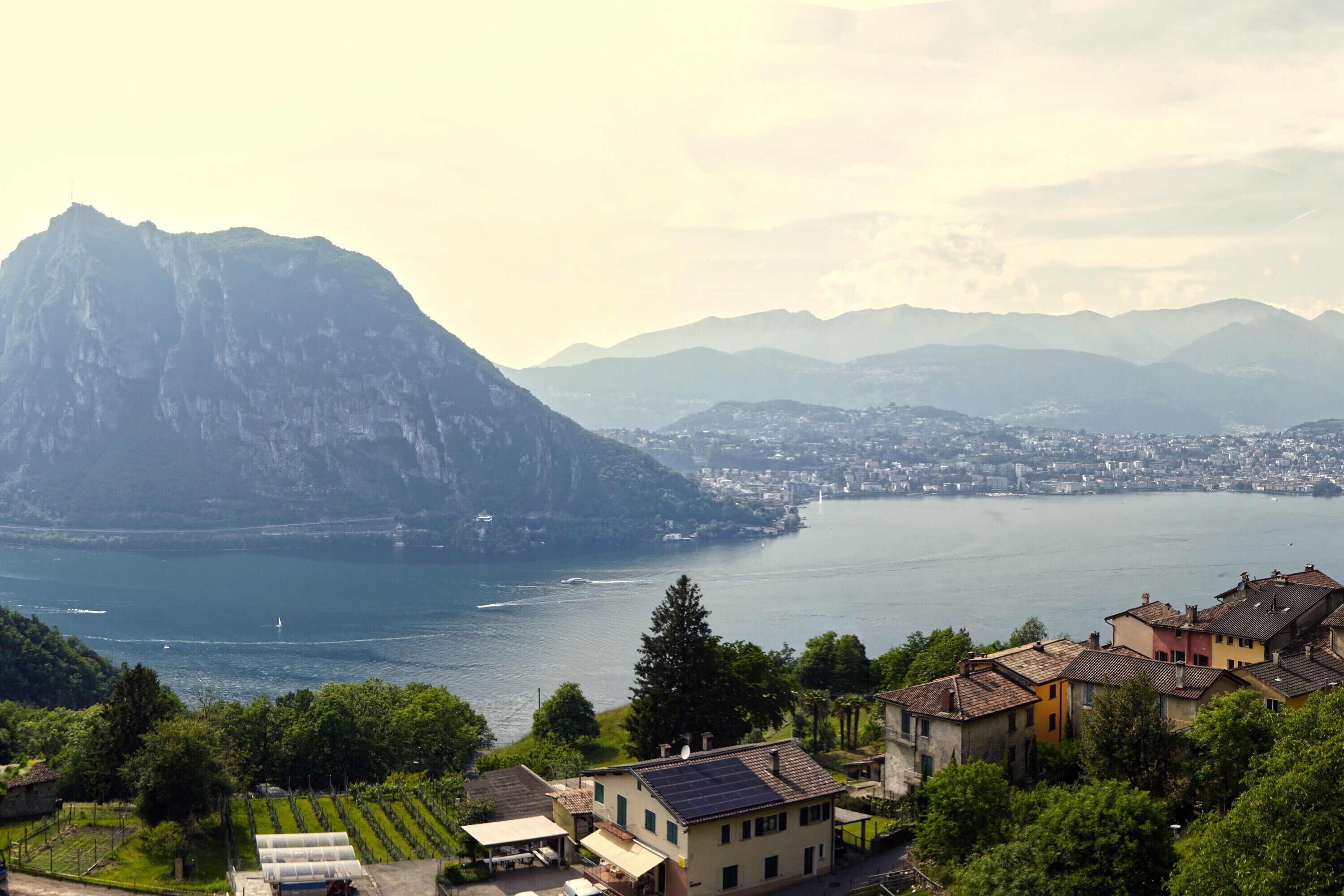 A lakeside town with a mountain backdrop, houses with solar panels, and a vineyard.