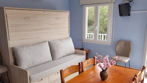 Ein Zimmer mit einer Holzdavonia, einem Esstisch mit Stühlen, einem an der Wand befestigten Fernseher und einem Fenster mit Blick auf Bäume.