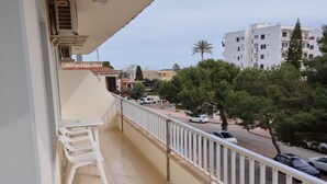 Ein Balkon mit weißer Brüstung, Tisch und Stühlen. Im Hintergrund sind Gebäude und Bäume zu sehen.