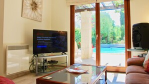 A living room with a flat-screen TV, a glass coffee table, a sofa, and a view of a swimming pool through large sliding glass doors. - Villa HG28 - Loukia (Kouklia)