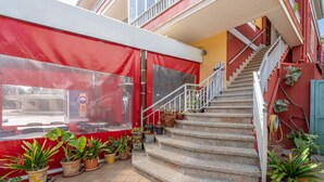 Una escalera lleva a un balcón con plantas en macetas y un toldo rojo.