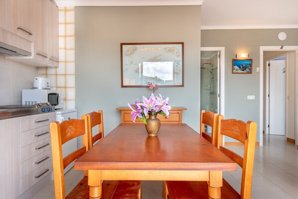 A dining area with a wooden table, chairs, a vase with flowers, a kitchen with appliances, and a framed map on the wall.