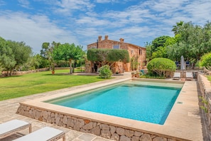Une piscine bordée de pierres, un mur de pierres et un bâtiment avec un jardin en arrière-plan.