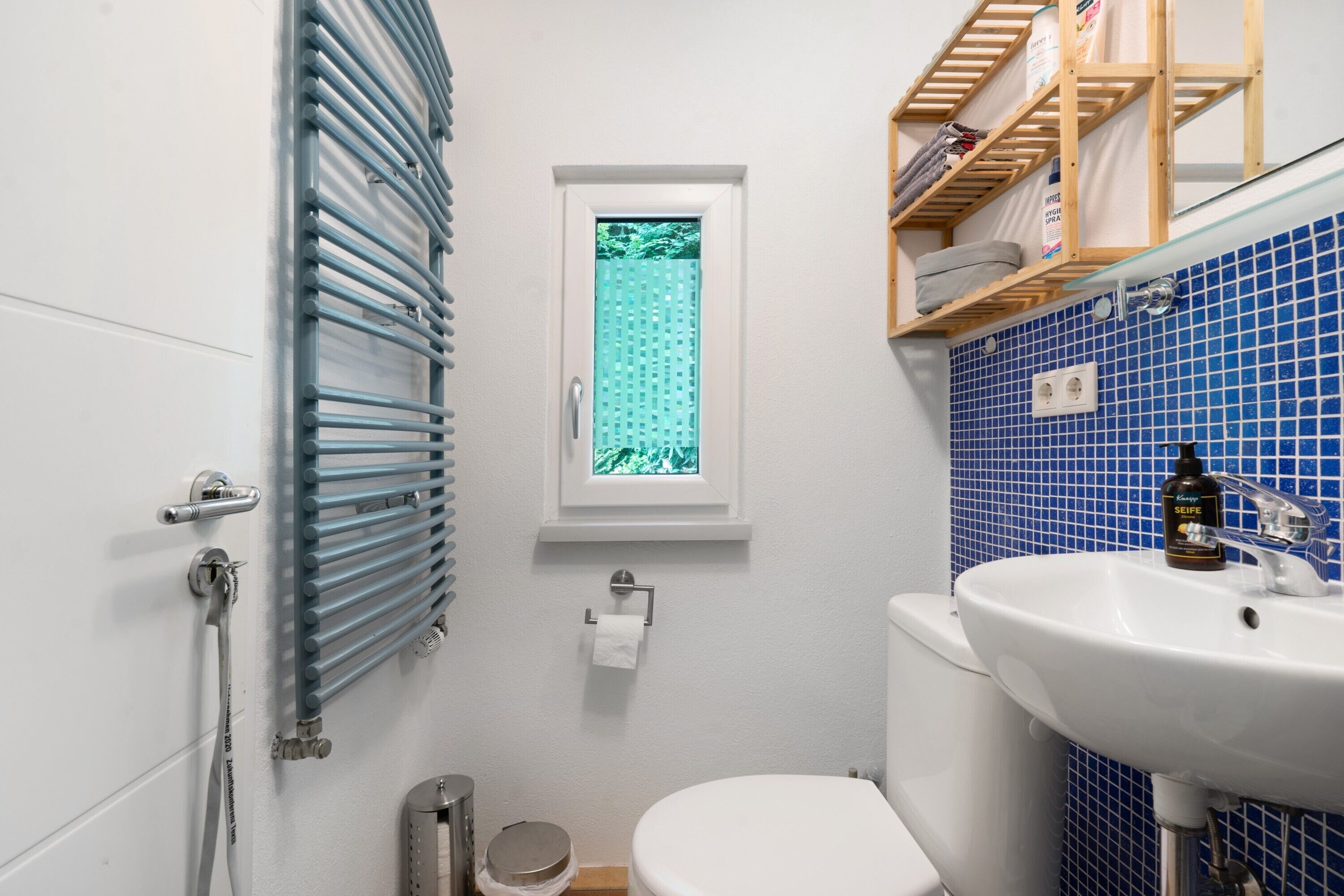 Ein Badezimmer mit Toilette, blauen Fliesen, einem Fenster mit Aussicht, einem Waschbecken und einem Handtuchhalter.
