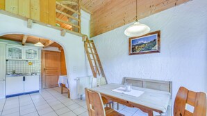Un interior rústico con una mesa de comedor de madera, sillas, una escalera, una cocina con ventana y un cuadro en la pared.
