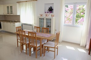 Ruokailutila, jossa on puinen pöytä ja tuolit, valkoisilla kaapeilla varustettu keittiö sekä ikkuna, jonka ulkopuolella on vaaleanpunaisia kukkia.