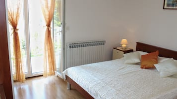 Un dormitorio con una cama, una mesita de noche con una lámpara, un radiador y cortinas translúcidas en una puerta corrediza de vidrio.