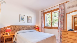 A bedroom with a bed, a wooden headboard, a nightstand with a lamp, a dresser, a window with curtains, and two framed pictures on the wall.