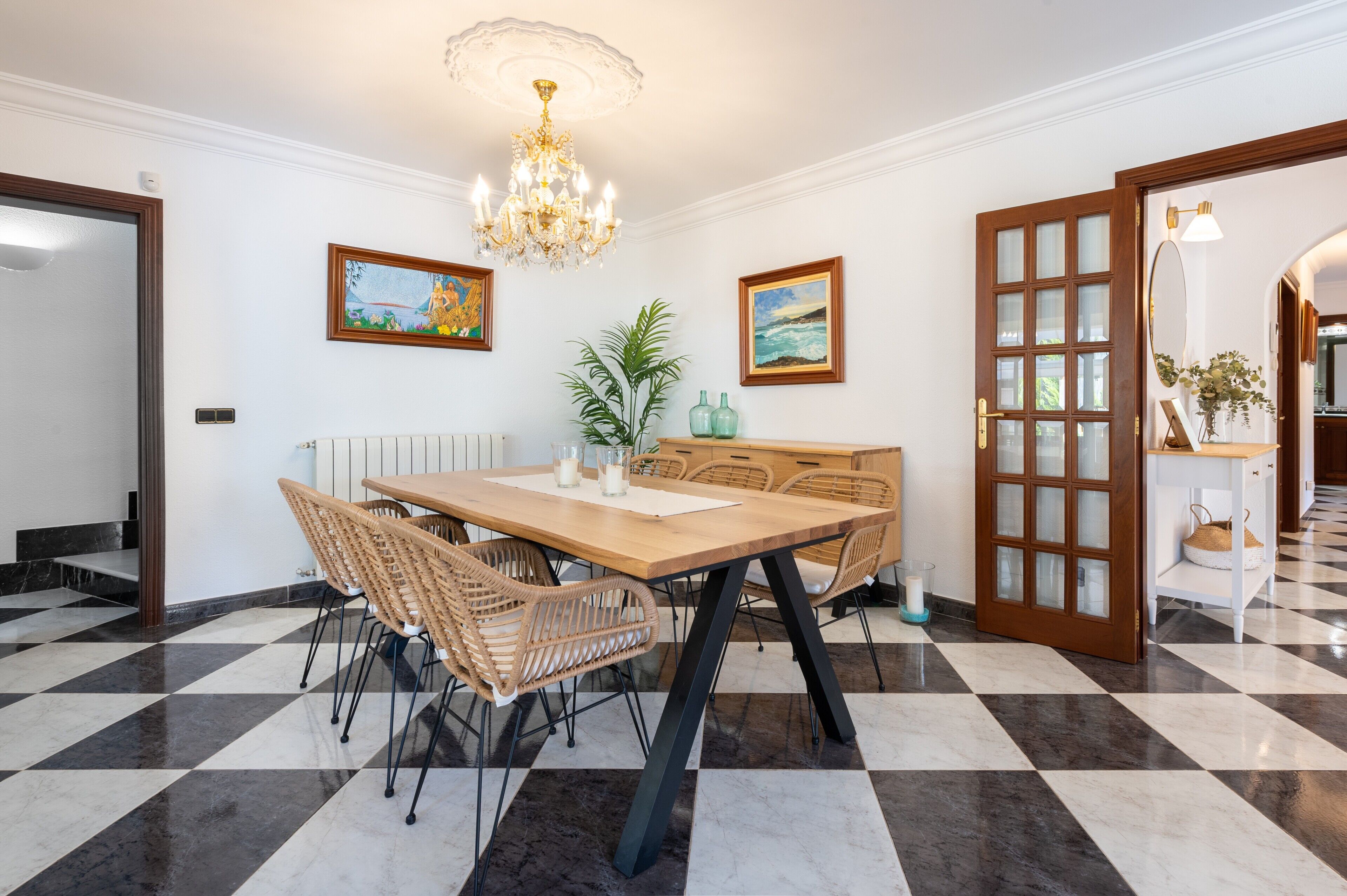 A dining area with a checkered floor, a chandelier, wicker chairs, a wooden table, and framed artwork on the walls.