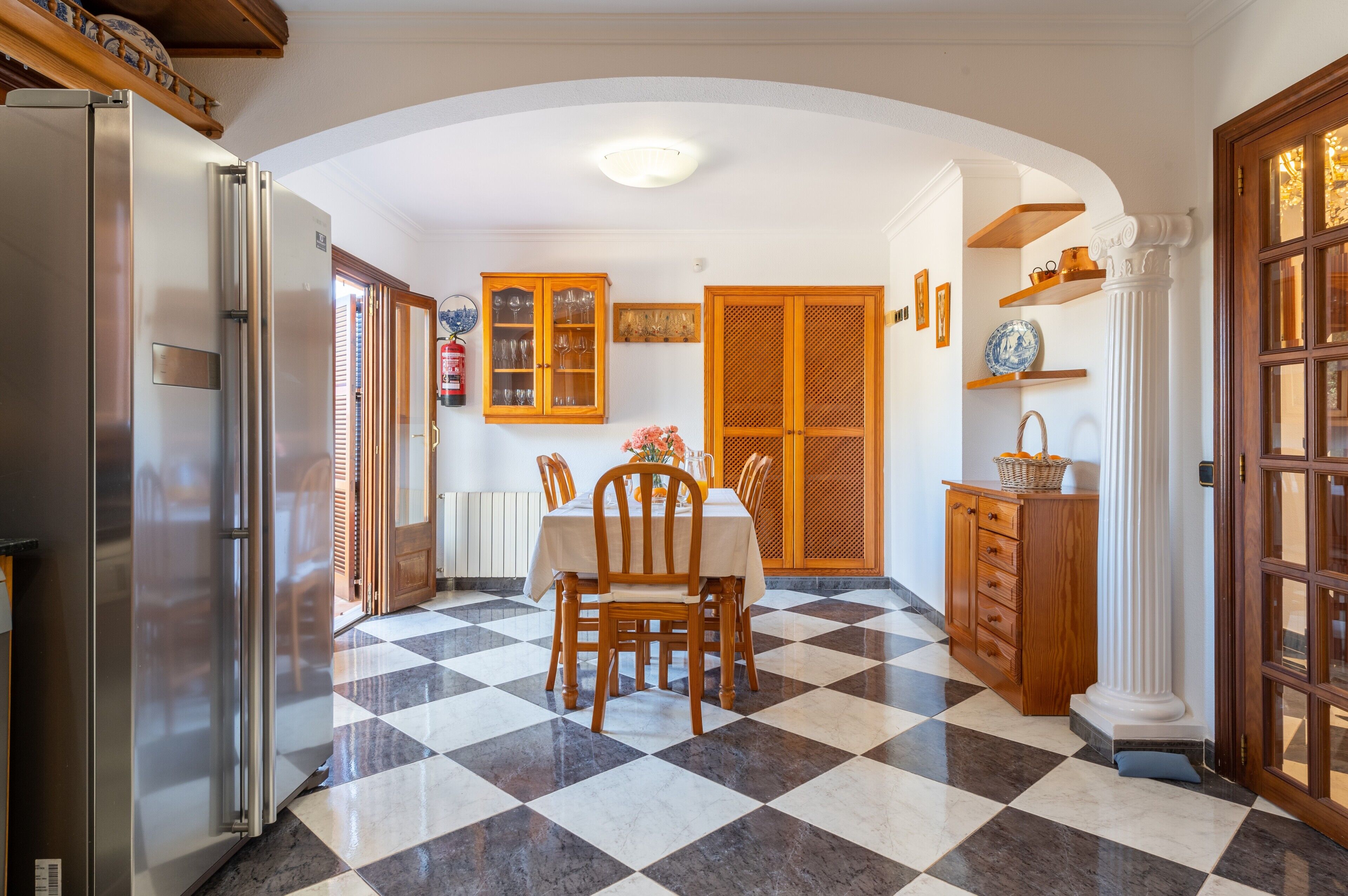 A kitchen with a checkered floor, a dining table with chairs, a refrigerator, wooden cabinets, and a column.