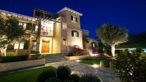 A two-story house with a stone facade, a wooden door, and a well-lit front yard with a lawn and trees. - Villa 141 - Rio (Kouklia)