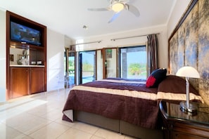 A spacious bedroom with a large bed, a ceiling fan, a TV mounted on the wall, and a view of the outdoors through the sliding glass doors.