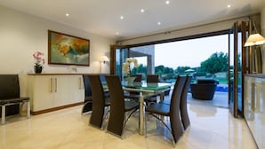 A modern dining area with a glass table and chairs, a painting on the wall, and a view of greenery outside. - Villa 141 - Rio (Kouklia)