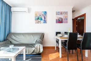 A living room with a sofa, a dining table with chairs, and two wall-mounted air conditioners.