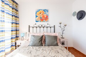 Un lit avec une literie à motifs floraux, un rideau à rayures, un tableau accroché au mur, une lampe de chevet et une petite table sur laquelle est posé un objet décoratif.