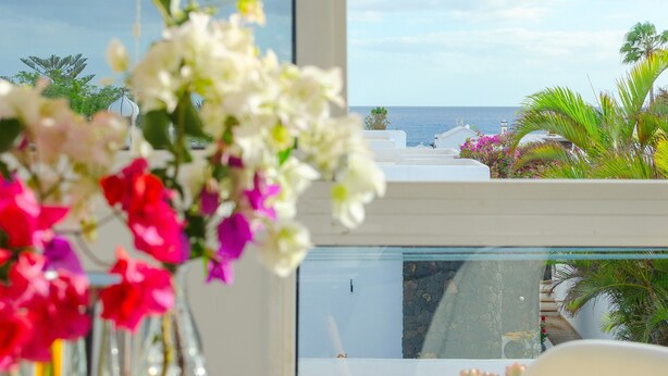 Un balcón con vista al mar, un edificio blanco y una barandilla de cristal.