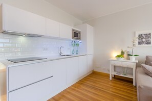 Moderni keittiö, jossa on valkoiset kaapit, mikroaaltouuni sekä pieni pöytä, jonka päällä on kasvi ja hedelmäkulho.