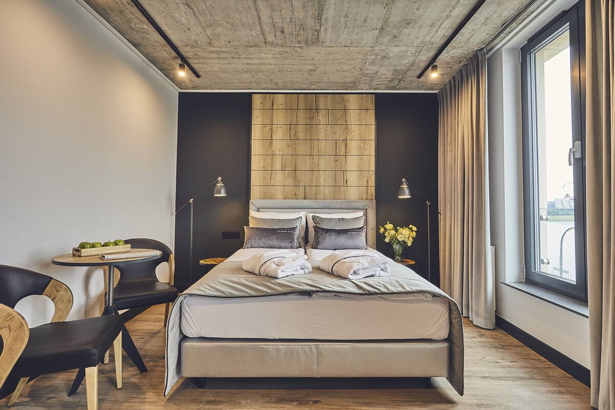 A modern bedroom with a large bed, a small table with a fruit bowl, two chairs, and a view of the city through a window.