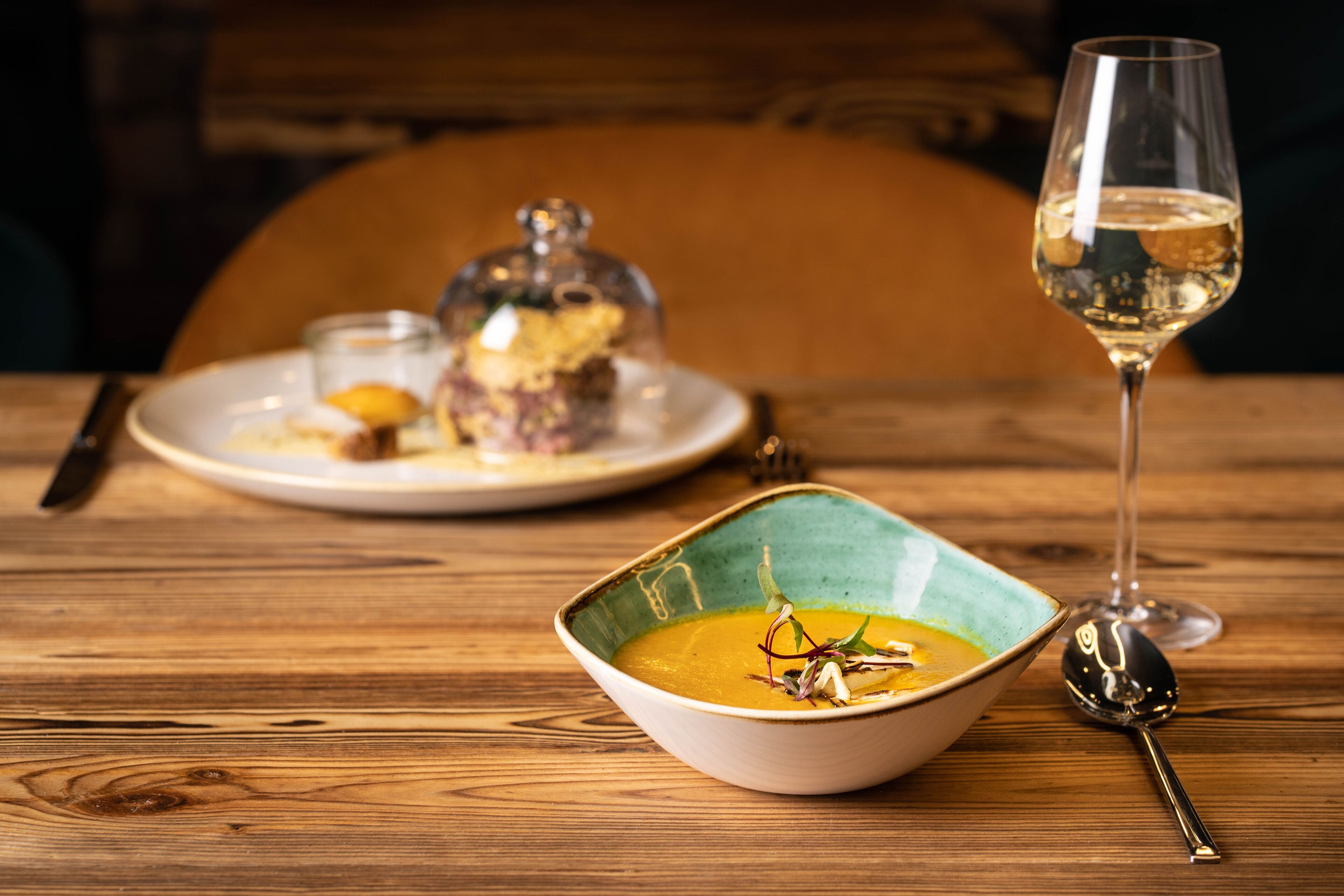 A wooden table with a bowl of soup, a glass of white wine, and a spoon.