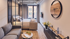 Camera d'albergo compatta con un letto, una poltrona, un tavolino con un libro e delle mele, un vaso con fiori e una sveglia a parete in legno.