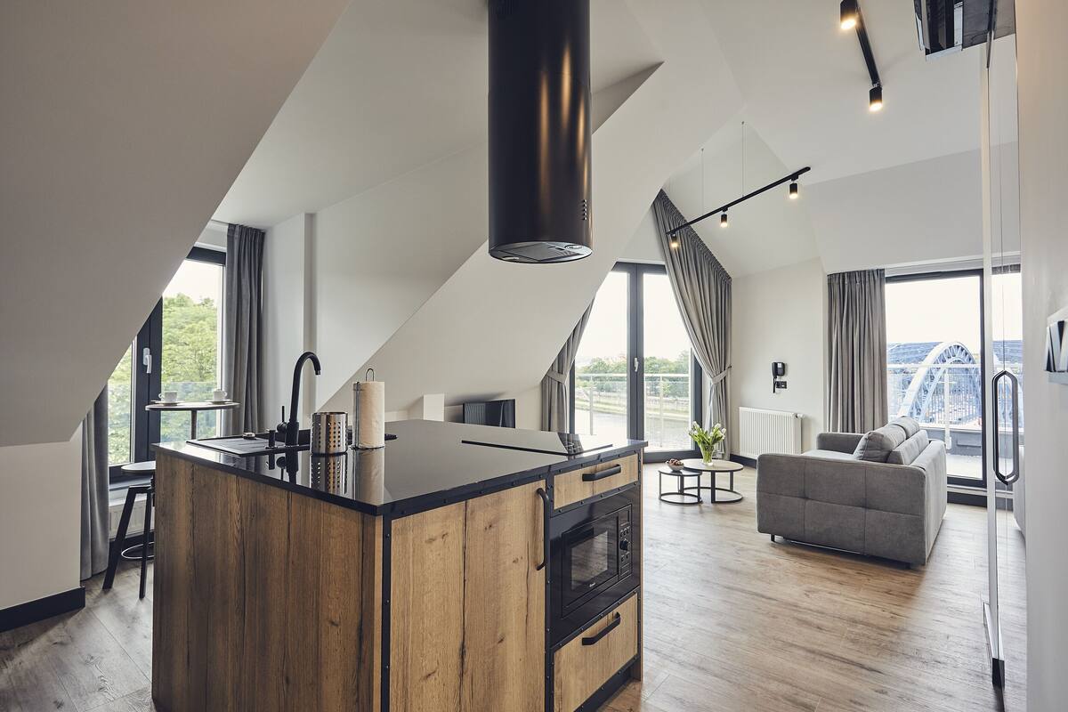 A modern kitchen with a wooden island, a dining area, and a living space with a sofa and a view of the outdoors.