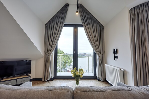 A modern living room with a large window, a flat-screen TV, a vase of flowers, and a sofa.