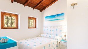 Un dormitorio con cama, dos ventanas, un cuadro, una mesita de noche y una lámpara.