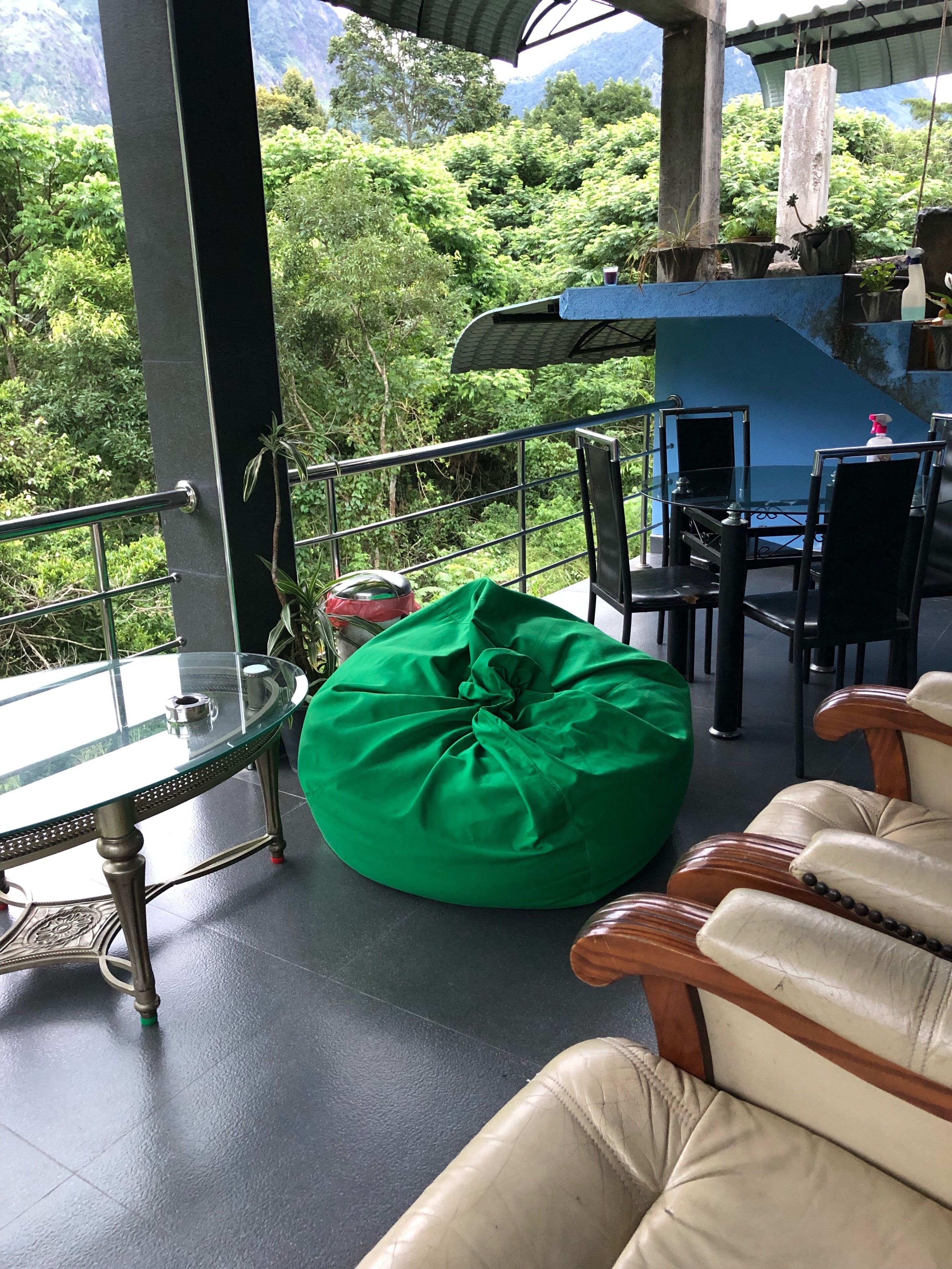 A balcony with a glass table, a green bean bag, leather chairs, and a view of lush greenery.