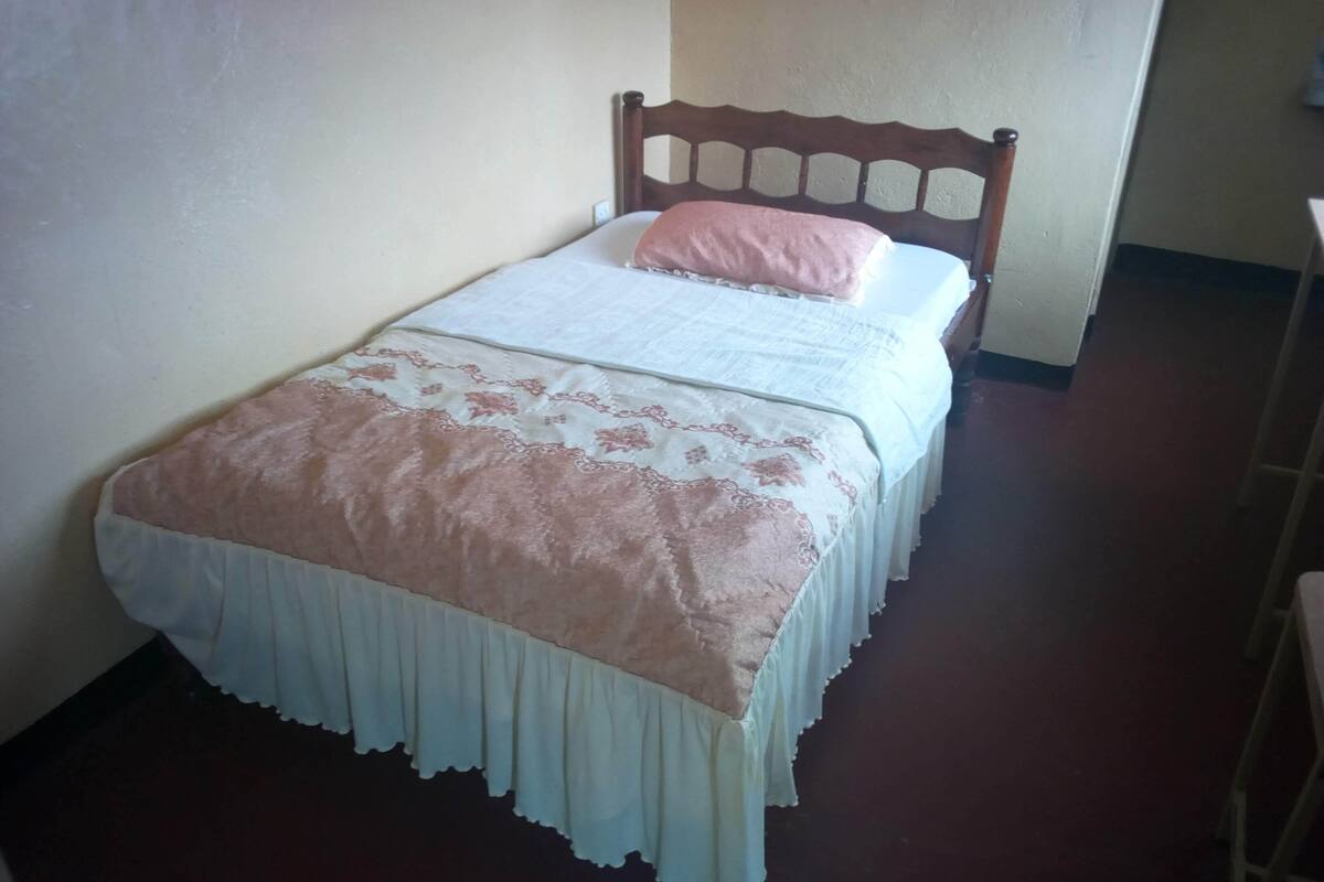A single bed with a wooden headboard, a white and pink bedspread, and a white skirted bed skirt.
