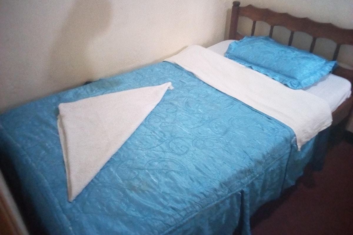 A single bed with a blue bedspread and white pillows in a room with plain walls.