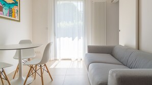 Uma sala de estar moderna com um sofá cinzento, uma mesa redonda com cadeiras e uma janela ampla com cortinas brancas.