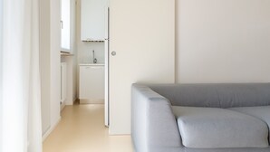 Quarto moderno e minimalista com sofá cinzento, armário branco e pequeno espaço de cozinha.