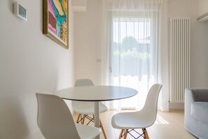 A modern dining area with a round table and white chairs, a painting on the wall, and a view through a large window.