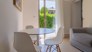 Uma sala de estar moderna com uma mesa redonda e cadeiras brancas, um sofá cinza e uma porta de vidro deslizante que dá para uma varanda com vegetação lá fora.