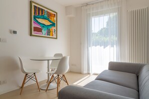 A modern living room with a grey sofa, a dining table with white chairs, and a colorful abstract painting on the wall.