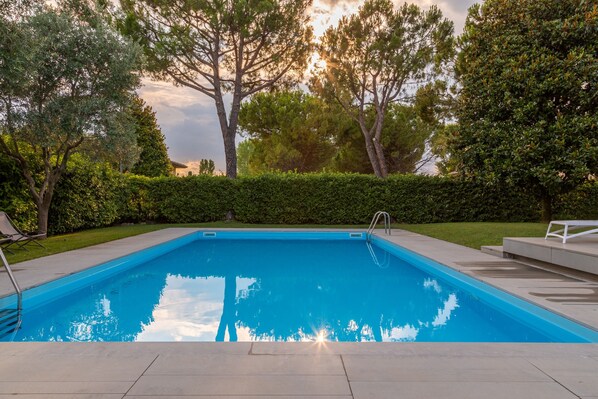 A rectangular swimming pool with a clear blue water surface, surrounded by a paved deck and a low hedge.