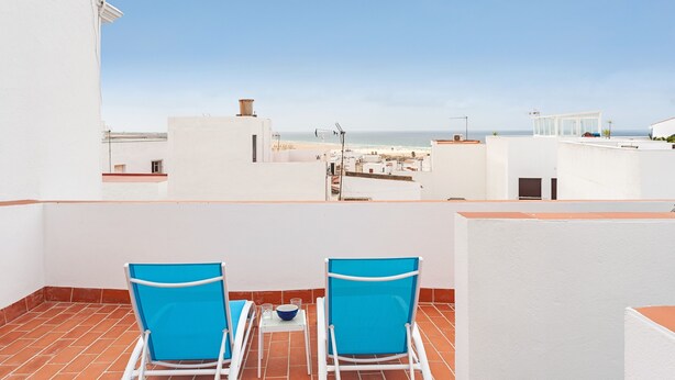 Een dakterras met twee blauwe stoelen, een kleine tafel en uitzicht op het strand en gebouwen.
