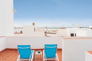 Una terraza en la azotea con dos sillas azules, una mesita y vistas a la playa y edificios.